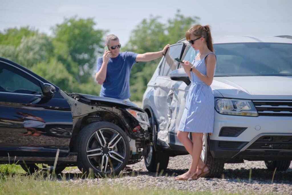 Woman in blue dress and man in jeans tshirt on phones after side collision car crash sunny day woods in background